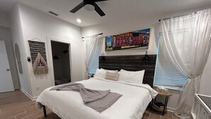 Un dormitorio moderno con una cama grande, un ventilador de techo, un cuadro en la pared y una ventana con cortinas.
