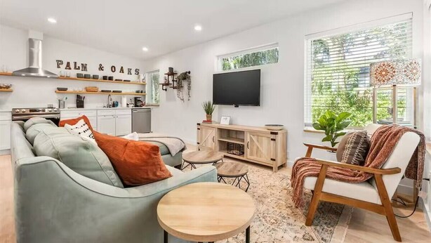 Una sala moderna con sofá, mesa de centro y sillón, una zona de cocina con estufa, y un televisor montado en la pared.