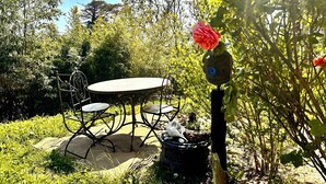 Un jardín con mesa y sillas de metal, rodeado de abundante vegetación y un comedero decorativo con una flor roja.