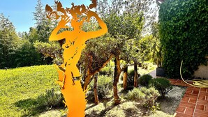 Un jardín con una gran escultura anaranjada de un árbol, rodeado de diversas plantas y un sendero de ladrillo.