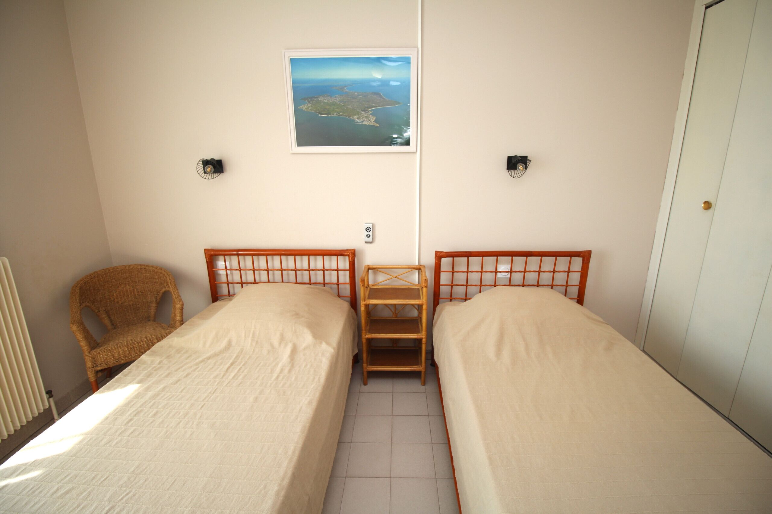 A small, clean room with two single beds, a wooden shelf, a wicker chair, and a framed picture on the wall.