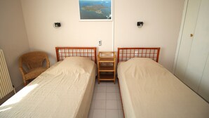 Una habitación pequeña y limpia con dos camas individuales, un estante de madera, una silla de mimbre y un cuadro en la pared.
