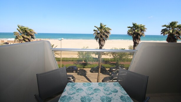 Un balcón con mesa y sillas que da a una playa con palmeras y cielo despejado.