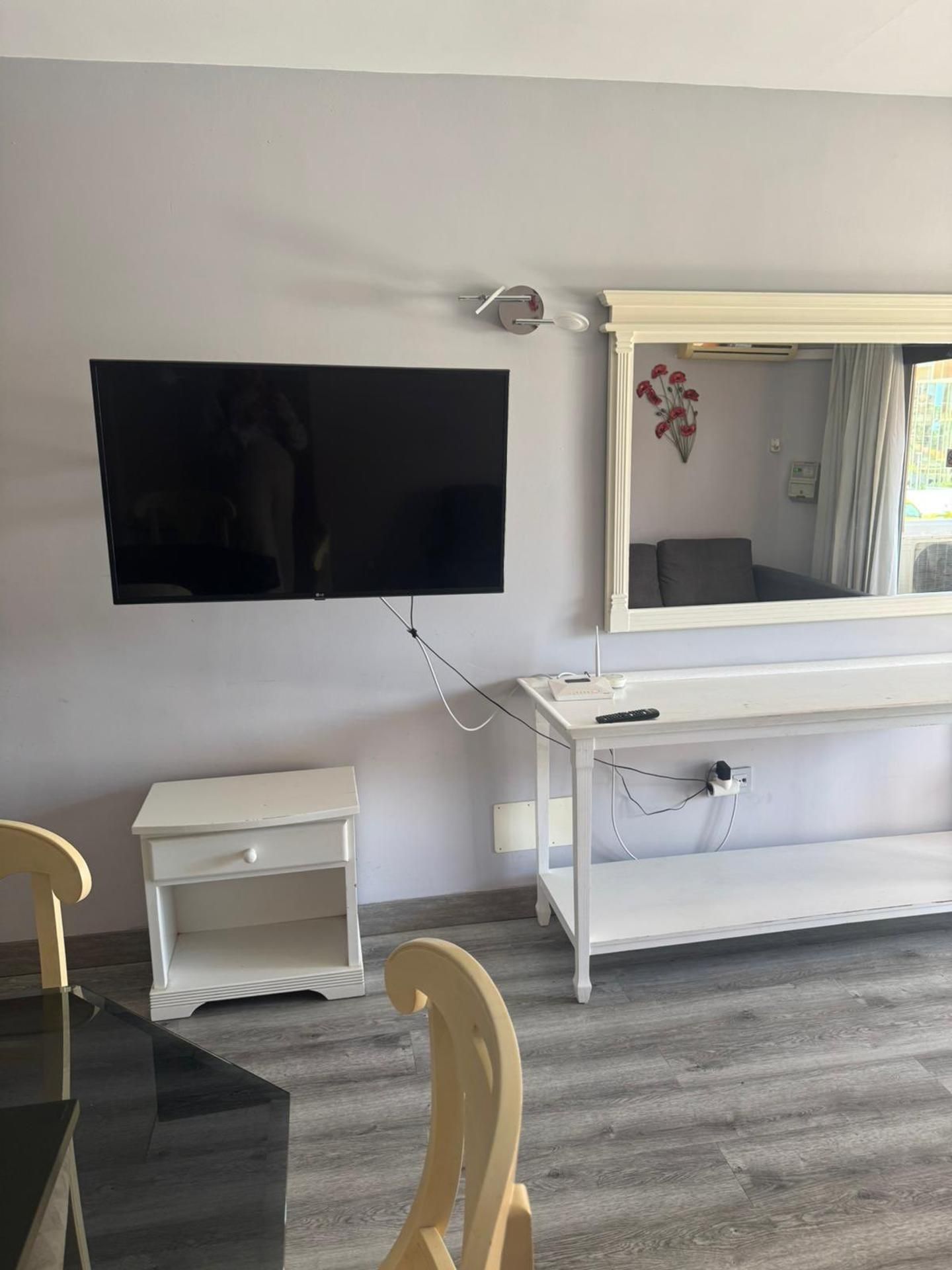 A room with a flat-screen TV mounted on the wall, a white console table, a small white drawer unit, a wooden chair, and a wooden dining table.