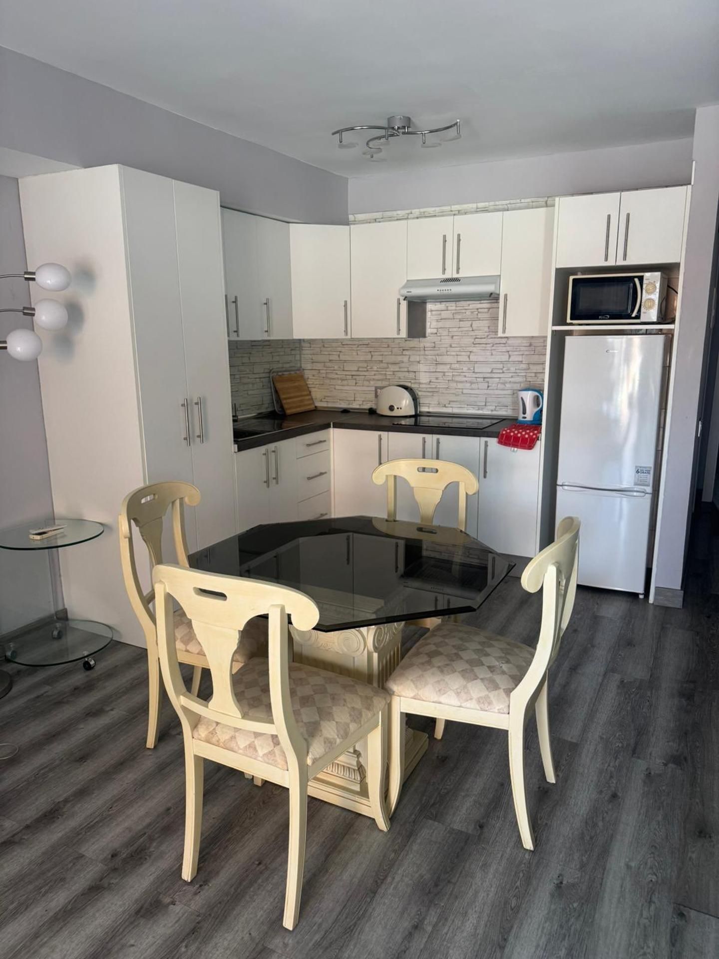 A compact kitchen with white cabinets, a black countertop, a microwave, and a refrigerator. There is a glass table with chairs.