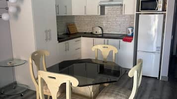 A compact kitchen with white cabinets, a black countertop, a microwave, and a refrigerator. There is a glass table with chairs.