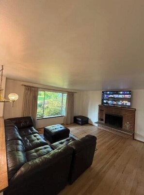 Relax in this inviting living room with a large sofa, flat-screen TV, and fireplace, surrounded by a view of lush greenery.