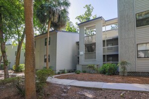 A multi-story residential building with a covered balcony on the second floor, surrounded by trees and shrubs.