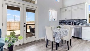 A modern kitchen with white cabinets, a dining area with a table and chairs, and a sliding glass door leading to an outdoor patio. - The Blue Flower (Rockport)