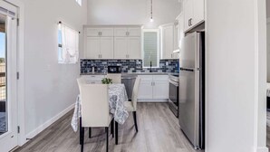 A modern kitchen with white cabinets, a stainless steel refrigerator, and a dining area with a table and chairs. - The Blue Flower (Rockport)