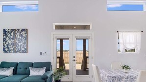 A living room with a sofa, a dining table with chairs, a potted plant, and a sliding glass door leading to an outdoor area. - The Blue Flower (Rockport)