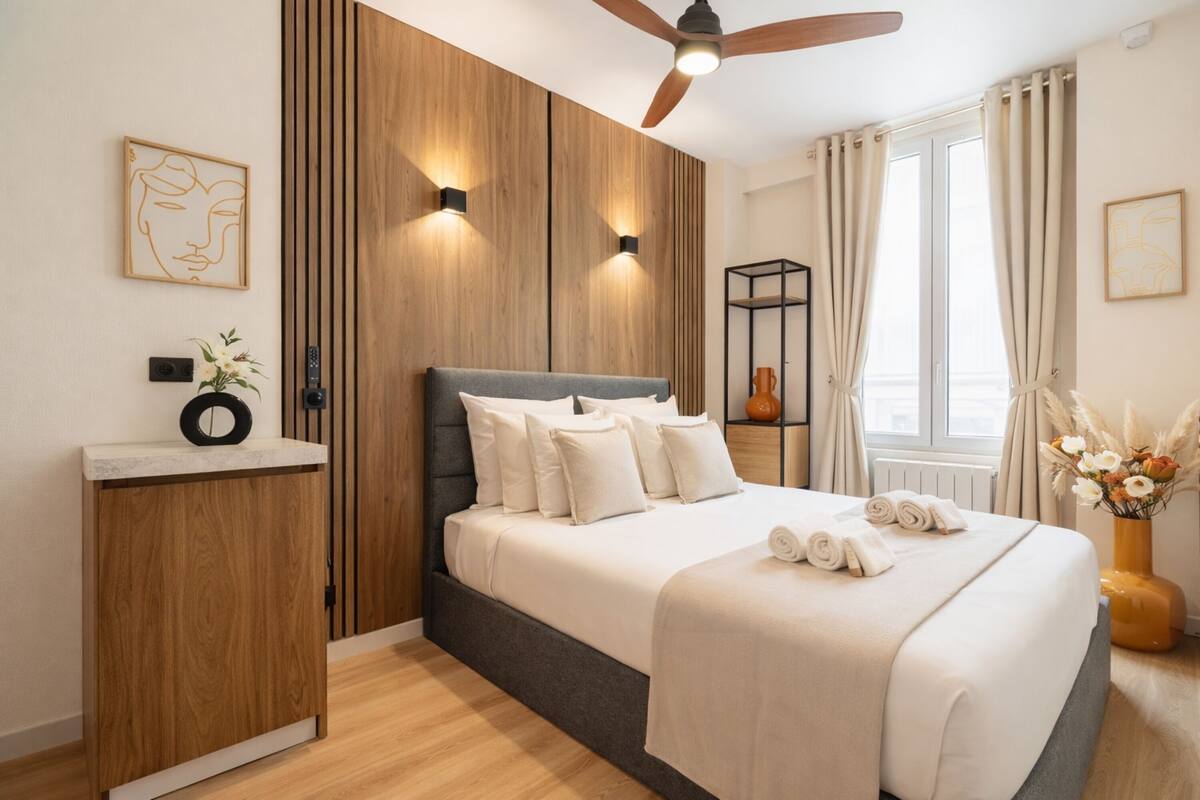 A modern bedroom with a bed, wooden paneling, a ceiling fan, and a window with curtains.
