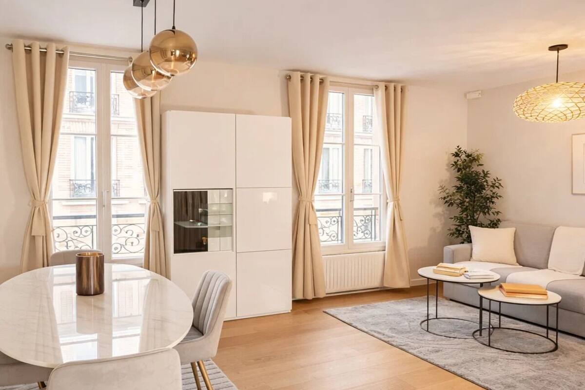 A modern living room with a round dining table, a sofa, and a coffee table. There are curtains, a plant, and a hanging light fixture.