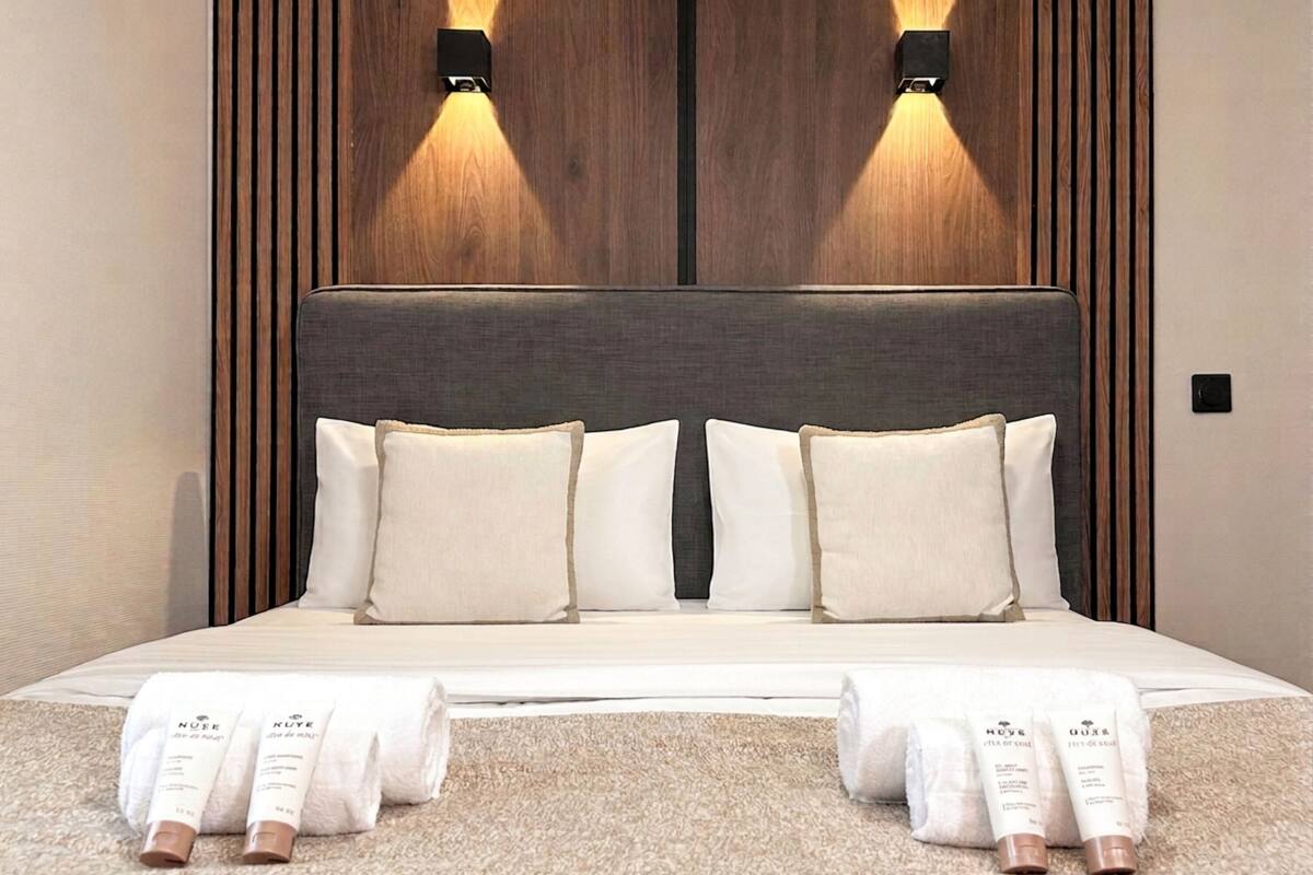 A neatly made bed with white bedding, two wall-mounted light fixtures, and a pair of white towels with brown caps on a beige carpet.