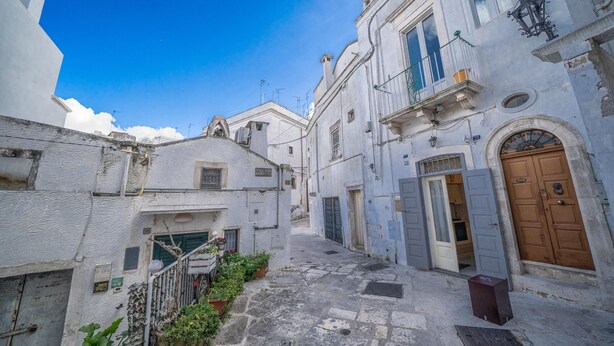 A narrow street with white buildings, a wooden door, and a small trash bin.