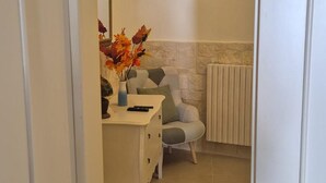 A narrow hallway with a white door on the left, a white dresser with a vase of flowers, a gray armchair, and a radiator on the right.