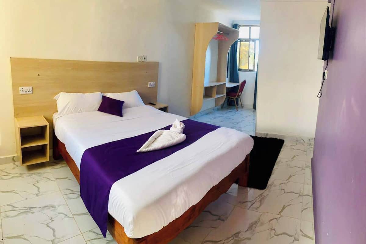 Una habitación de hotel con una cama, cabecera de madera, un pequeño estante de madera, una silla y un televisor montado en la pared.
