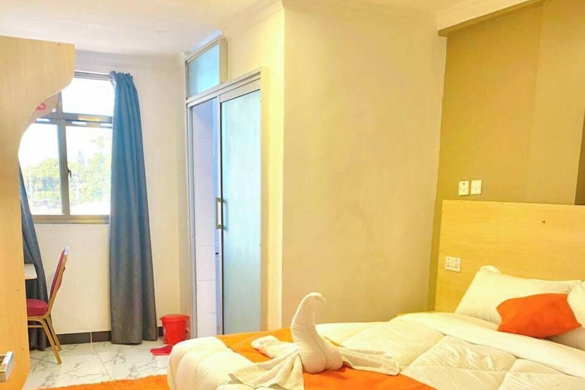 Una habitación de hotel con una cama, cabecera de madera, ventana con cortinas, una puerta y una pequeña mesa con una silla.