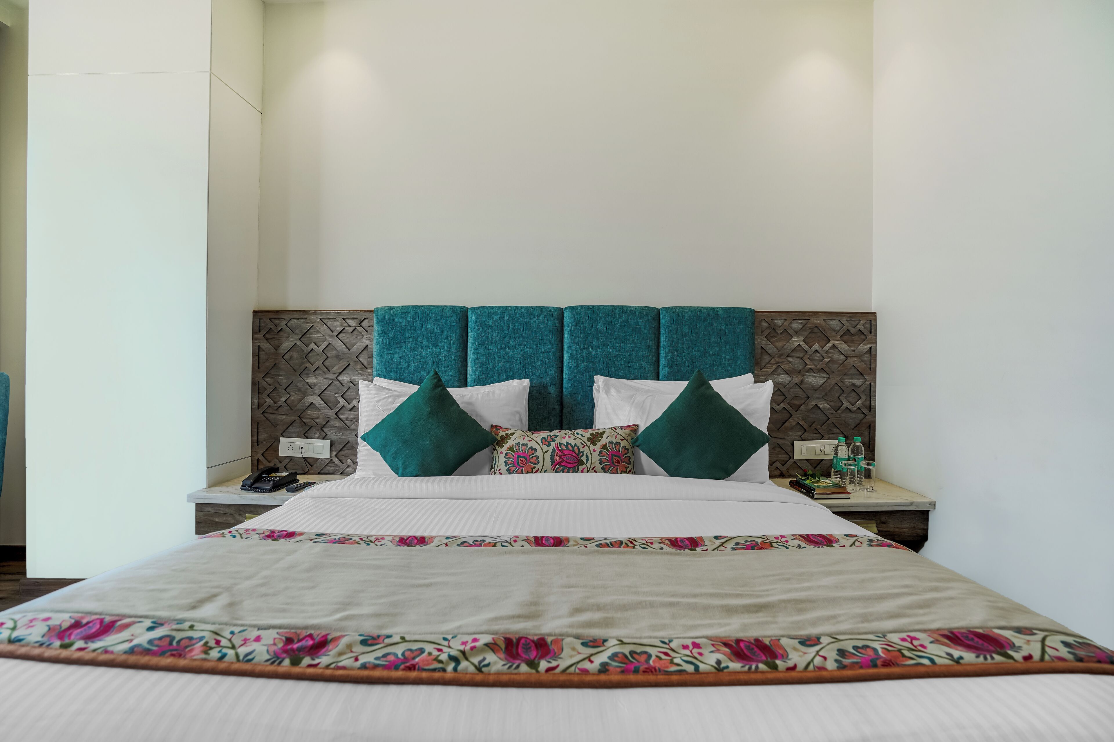 A neatly made bed with floral bedspread, a headboard with a geometric pattern, and two bedside tables with water bottles and a phone.