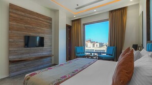 A hotel room with a large bed, a flat-screen TV mounted on a wooden panel wall, two blue chairs, a small table, and a large window with a city view. - A K CONTINENTAL (Amritsar)