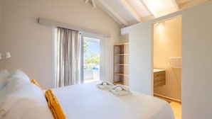 A hotel room with a large bed, a window with curtains, a wooden shelf, and a bathroom visible through an open door. - Ser na Private Retreat (St. Marie)