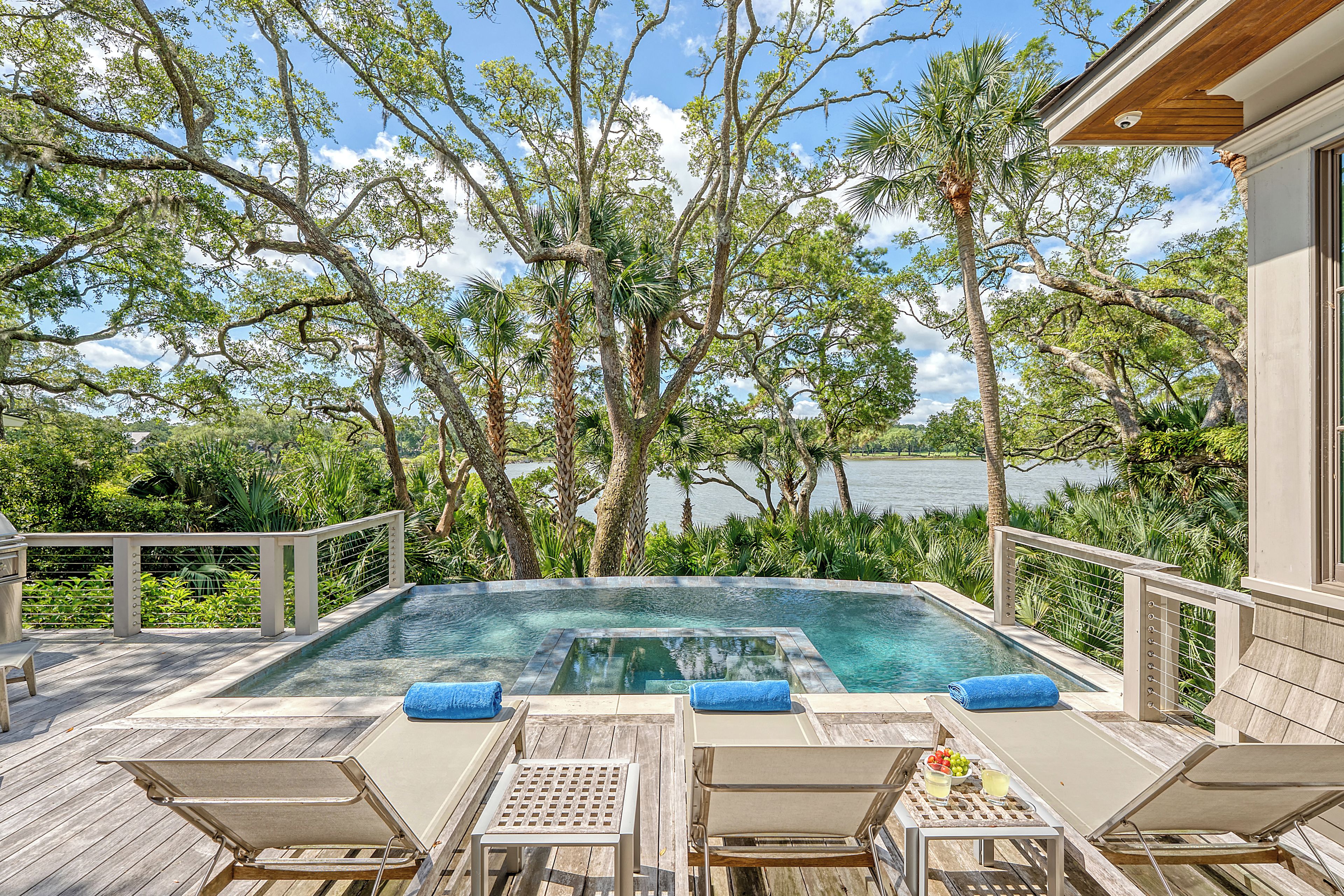 Une piscine à débordement avec des chaises longues et une table, entourée d’arbres et offrant une vue sur l’eau.