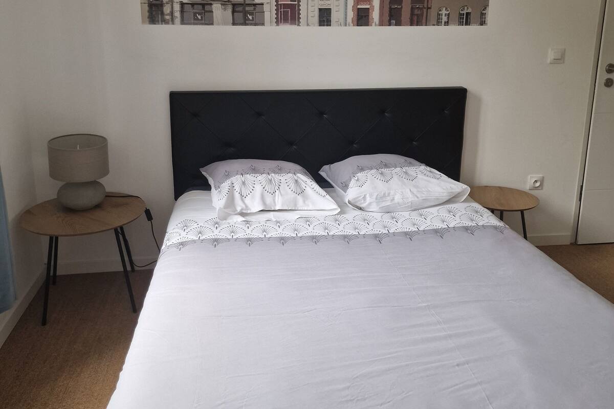 A neatly made bed with white linens and lace pillows, a round wooden side table with a lamp, and a wall-mounted painting of European architecture.