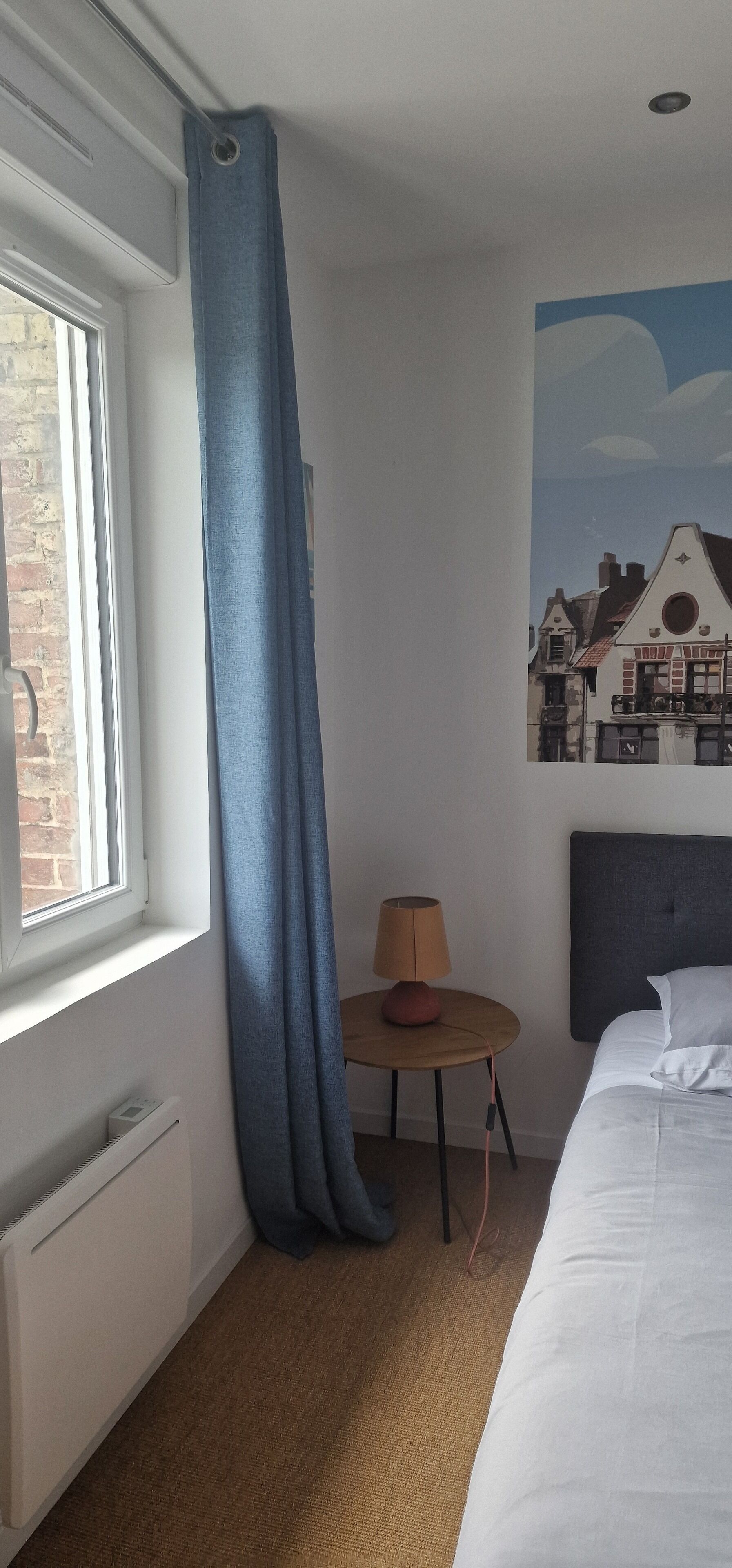 Une chambre d’hôtel avec un lit, une petite table avec une lampe, une fenêtre avec un rideau et un tableau au mur.