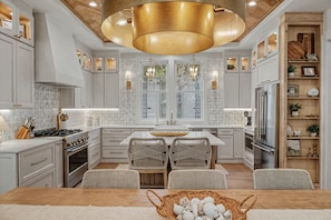 A modern kitchen with white cabinetry, a central island, wicker chairs, and a large chandelier.