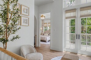 A room with a wooden floor, a large window with a view of greenery, a bed with a white headboard, and a plant on the left.