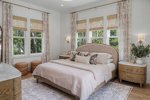 A bedroom with a large bed, two bedside tables, a nightstand, a marble bedside table, and a window with curtains.