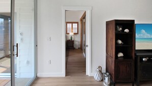 A hallway with a wooden floor, a sliding glass door, a dark wooden cabinet with decorative items, and a painting of a beach scene.