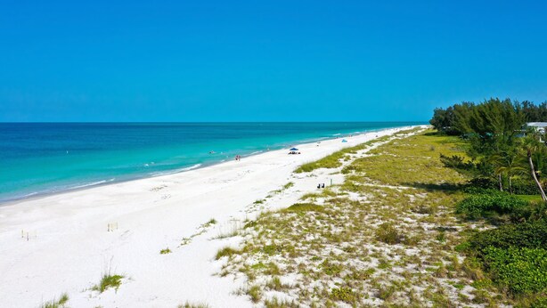 A beach with white sand, clear turquoise water, and a few people. There are some trees and a building in the background.