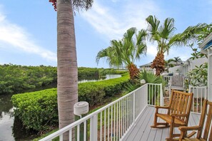 Um deck com cadeiras de madeira, um corrimão branco e vista para um rio e vegetação.