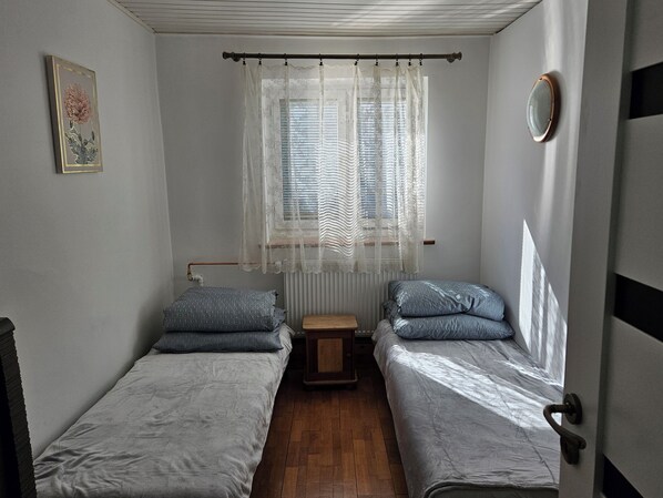 Un alojamiento pequeño y con una sola habitación, con dos camas individuales, un radiador, una mesita y una ventana con cortinas traslúcidas.
