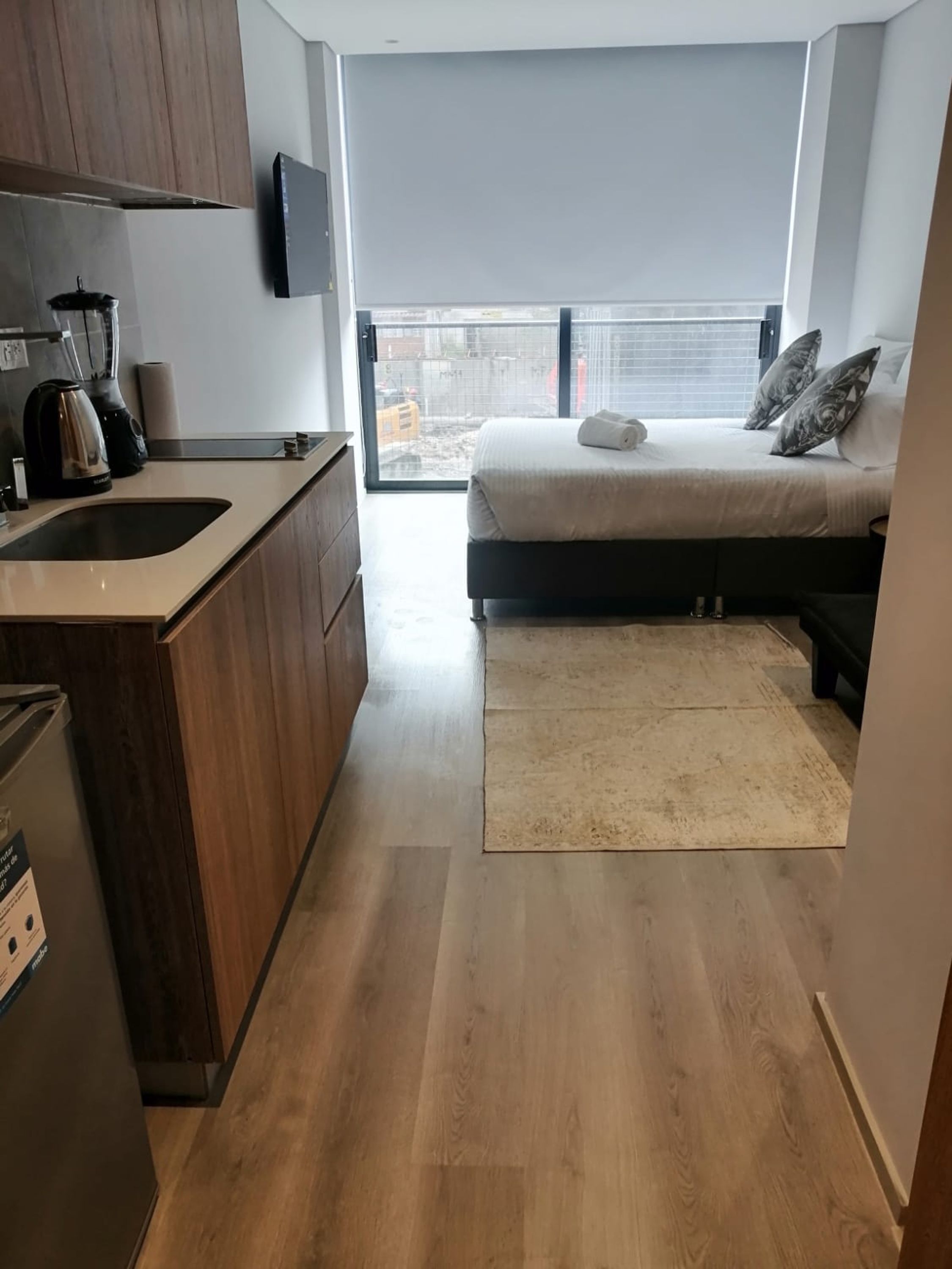 A compact kitchen with a built-in stove, a sink, and a coffee maker. There is a bed with a white mattress and grey pillows, and a large window providing a view of the outside.