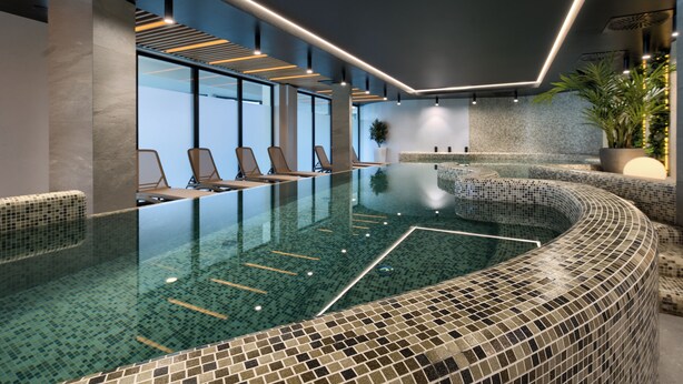 Una piscina interior moderna con diseño de mosaicos, sillones de descanso y vista al cielo.