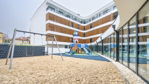 Un parque infantil con columpios y toboganes, rodeado por un edificio con varios balcones y ventanas amplias.