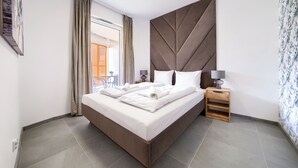 Una cama bien hecha con sábanas blancas, una mesita de noche de madera con lámpara, y vistas a un balcón exterior con mesa y sillas.