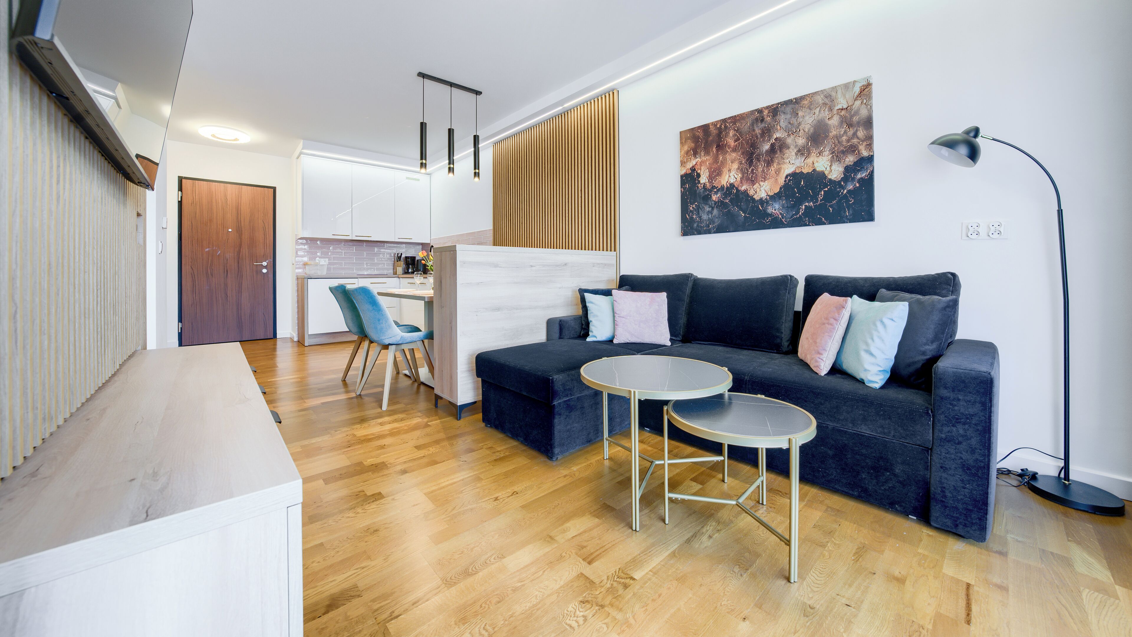 Una sala moderna con un sofá azul oscuro, una mesa de centro redonda y una silla azul. Se observa una cocina al fondo con armarios blancos y una encimera de madera.