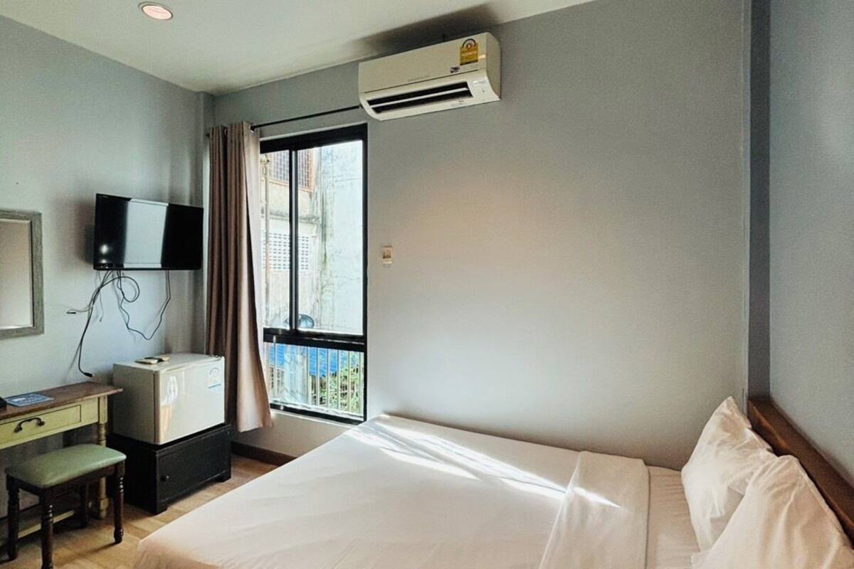 A hotel room with a large bed, a desk with a computer, a flat-screen TV, and an air conditioning unit.