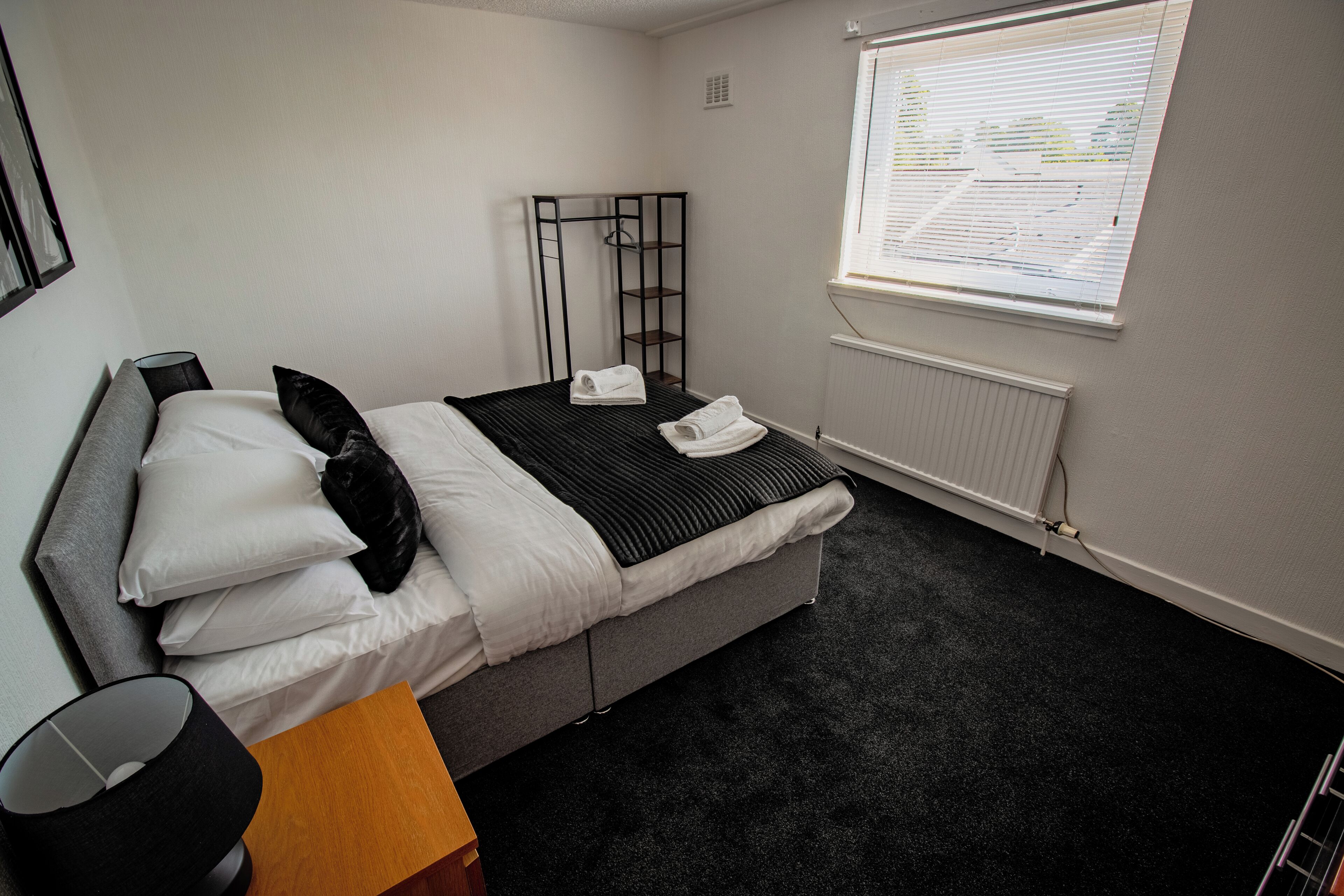 Una habitación de hotel moderna con una cama gris, ropa de cama blanca, una manta a rayas negras y blancas, una almohada negra, una lámpara negra, una mesita de noche de madera y una ventana con persianas.