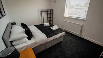 Una habitación de hotel moderna con una cama gris, ropa de cama blanca, una manta a rayas negras y blancas, una almohada negra, una lámpara negra, una mesita de noche de madera y una ventana con persianas.