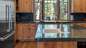 A kitchen with wooden cabinets, a stainless steel refrigerator, granite countertops, a large window with a view of trees, and a built-in sink. - GH261 | Amazing Luxury home sure to be your new favorite! (Black Butte Ranch)