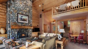 A cozy log cabin interior with a stone fireplace, a sofa set, a dining table, and a balcony with a wooden railing. - GH261 | Amazing Luxury home sure to be your new favorite! (Black Butte Ranch)