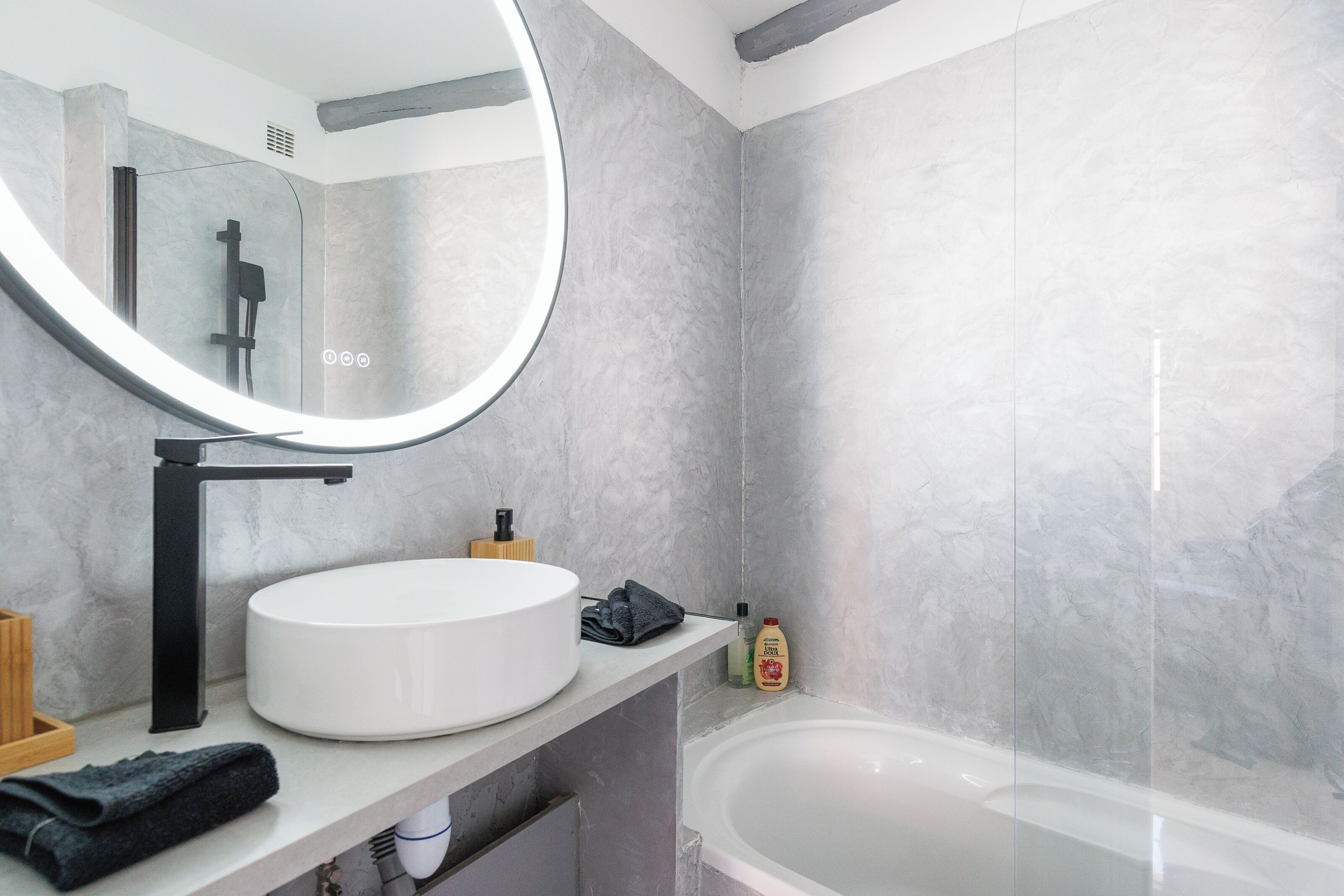 A bathroom with a round mirror, a white sink, a black faucet, a bathtub, and marble walls.