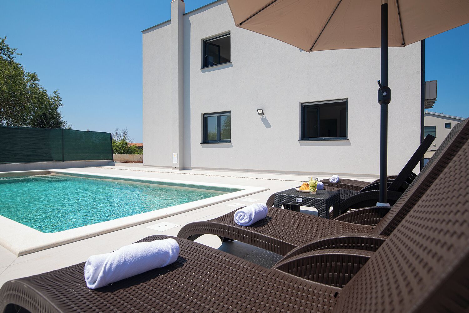Un espace piscine extérieur moderne, doté de chaises longues et d’un parasol, entouré d’un bâtiment blanc avec de nombreuses fenêtres.