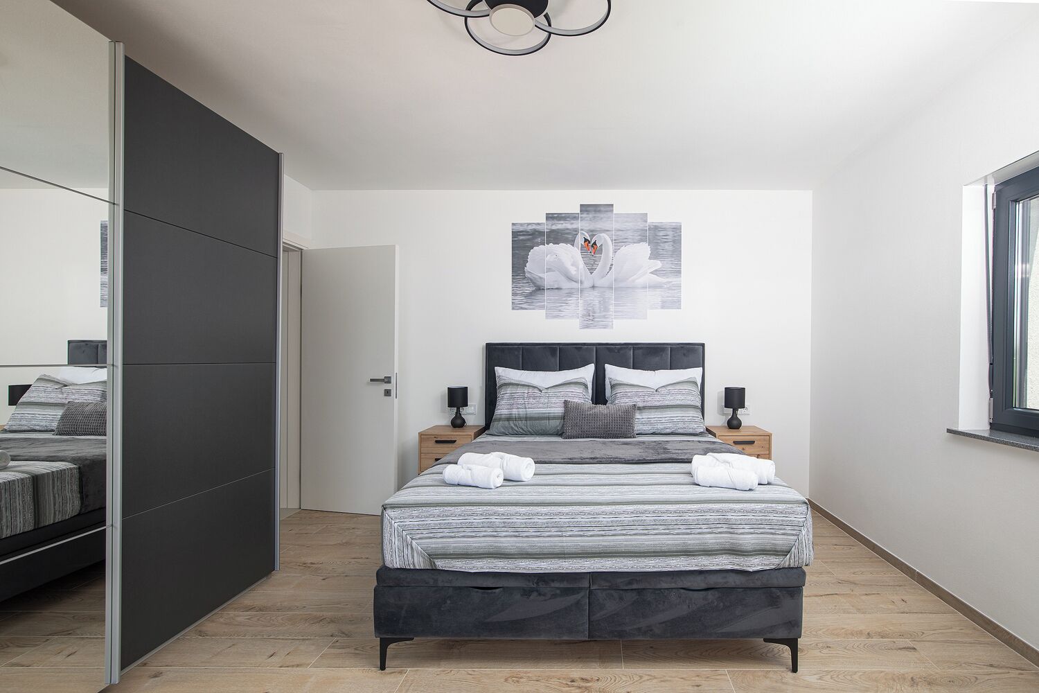 A modern bedroom with a large bed, a wardrobe, a bedside table, a nightstand, and a window.