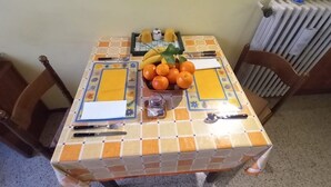 Une table à manger recouverte d’une nappe à motifs, un panier de fruits contenant des oranges et des bananes, et un petit récipient à sel.