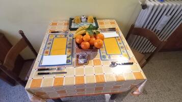 Un tavolo da pranzo con una tovaglia piastrellata, una ciotola di frutta con arance e banane, e un piccolo contenitore per il sale.